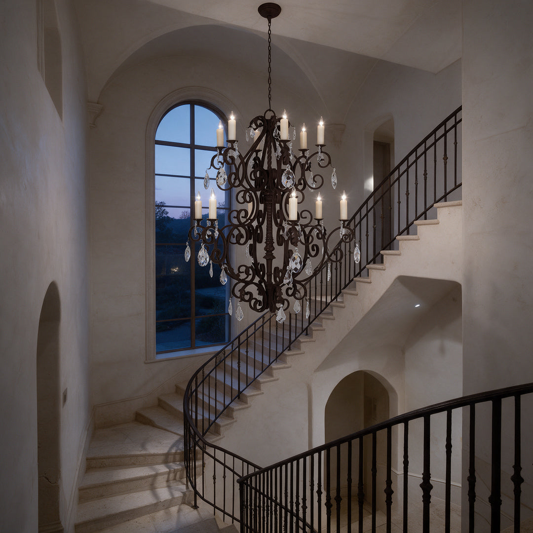 Monte Carlo Heritage 12-light crystal chandelier illuminated in a grand stair hall at twilight, beside an arched window and iron railing.