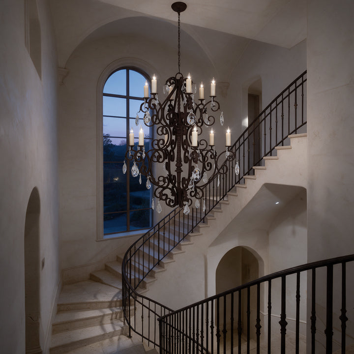 Monte Carlo Heritage 12-light crystal chandelier illuminated in a grand stair hall at twilight, beside an arched window and iron railing.