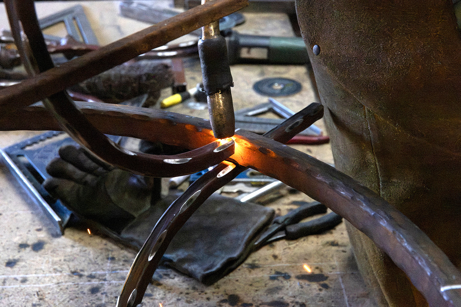 Ironworker welding curved iron arms together at the workbench during assembly of a handcrafted lighting fixture.